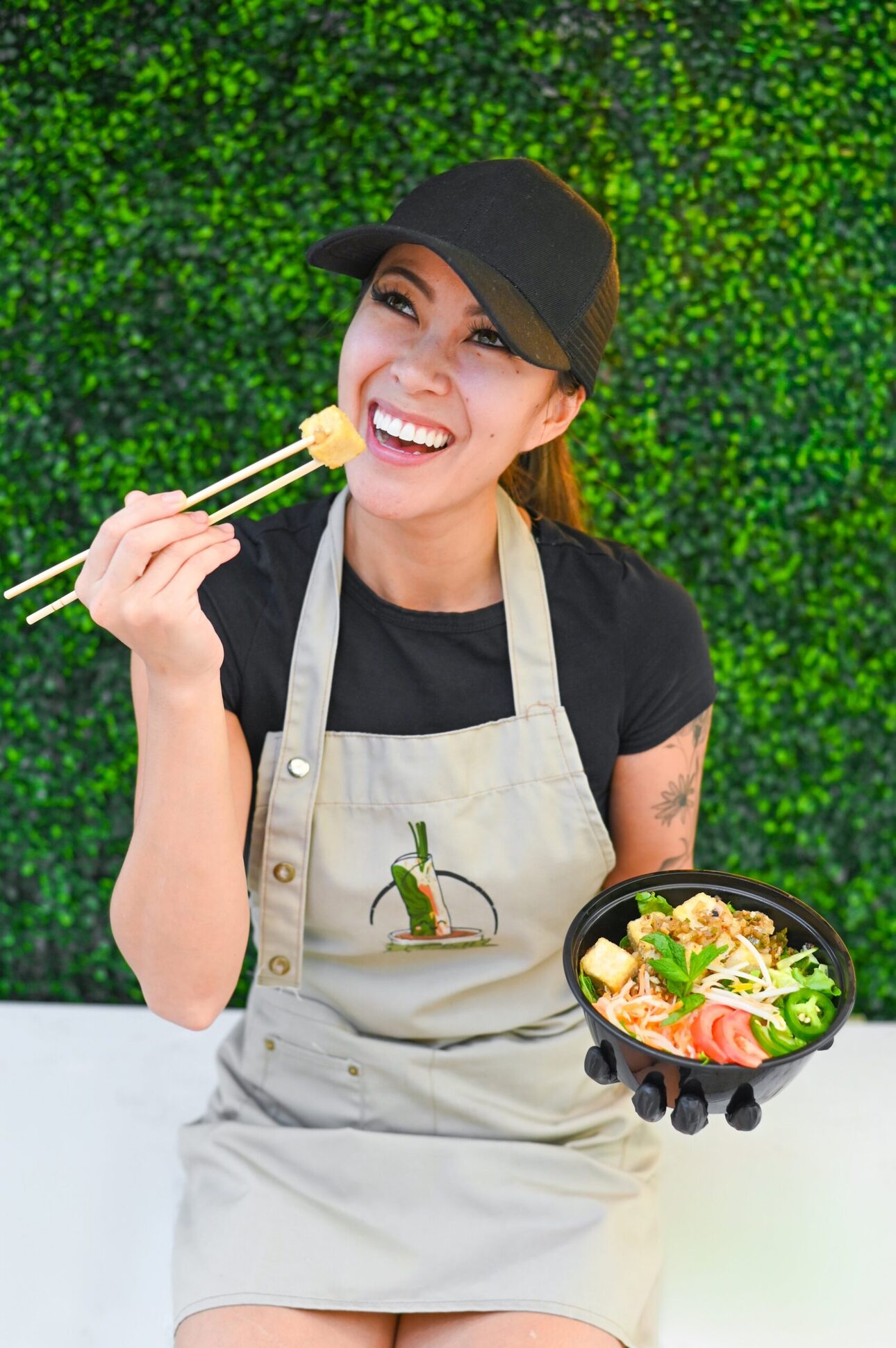 A V-Roll cook in a V-Roll apron lifting a bite with chopsticks, a noodle bowl in hand, in front of the greenery wall