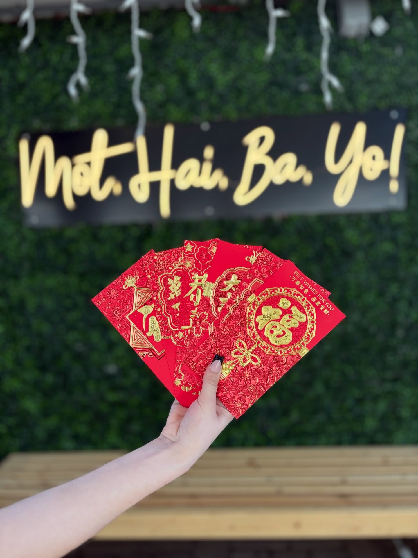 Red Lunar New Year envelopes held up under the Một, Hai, Ba, Yo! neon sign