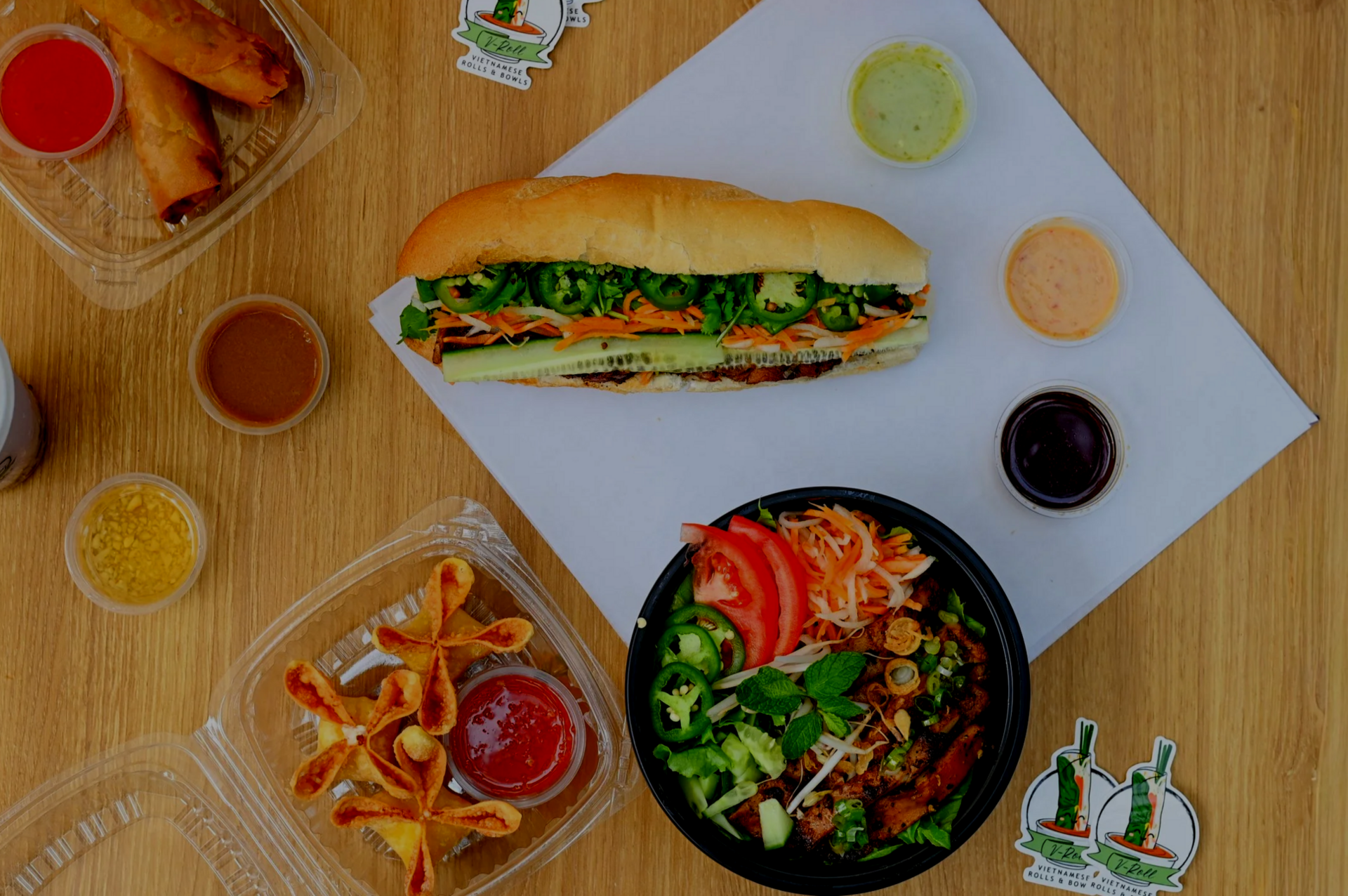 Overhead V-Roll spread of bánh mì, bowl, egg rolls and sauces on a wood table